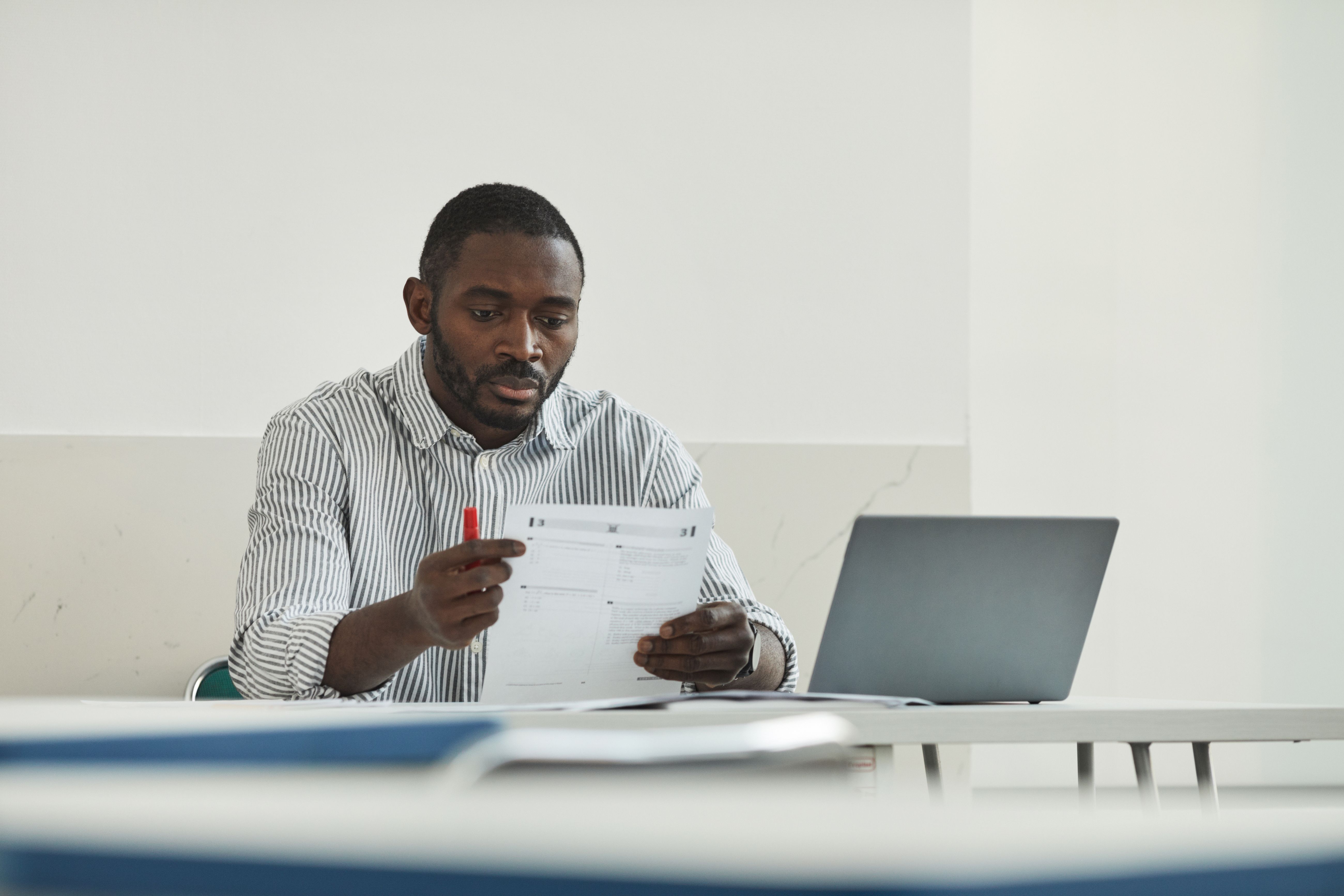 Teacher grading test papers at a workspace with a laptop, checking student work for AI use
